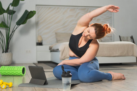 Yoga fitness workout training online. Woman practicing stretching with online app lessons at home. Plus size female doing sports exercise on yoga mat indoor. Girl training workout for weight lossの写真素材