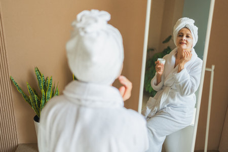 Mature adult old woman looking at mirror applying nourishing anti age face cream doing daily beauty routine. Senior woman with white mask creme on skin gets facial treatment Skin care aging skincareの写真素材