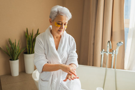 Mature adult old woman applying moisturizing body lotion on hands. Senior lady rubbing nourishing anti age cream doing morning daily skincare routine in bathroom. Beauty treatment spa body skin careの写真素材