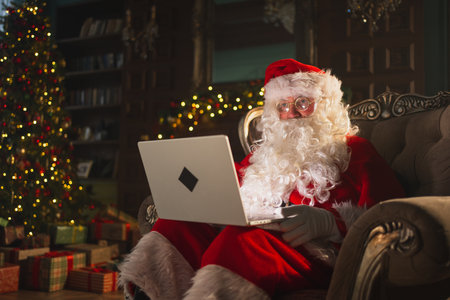 Merry Christmas. Santa Claus with laptop reading letters from children typing buying presents gifts online. Santa with computer on Christmas eve. Mail gifts of Santa Claus. Winter Holiday Celebrationsの写真素材