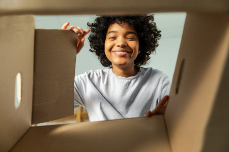 Inside of cardbox bottom view. African girl unpacking delivery looking in box. Woman opening carton box. Female getting parcel looking at delivered goods items. Satisfied client positive feedbackの写真素材