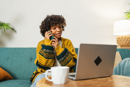 African American woman freelancer using laptop talking on phone at home office. Happy business girl having virtual meeting. Woman using phone laptop for communication working indoors. Remote workの写真素材