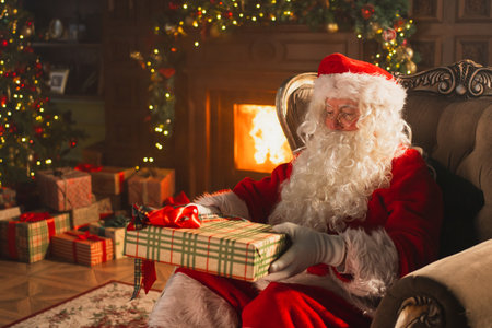 Merry Christmas. Santa Claus sitting in chair packing wrapping gift box in festive living room with fireplace decorations Christmas tree. Santa with gifts on Christmas eve. Winter Holiday Celebrationsの写真素材