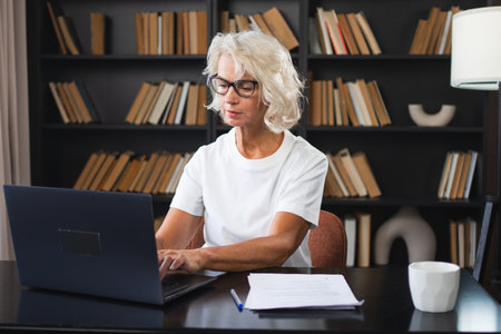 Confident stylish middle aged senior woman using laptop at workplace. Older mature lady businesswoman sitting at office table with laptop and paper document. Boss leader teacher professional workerの写真素材