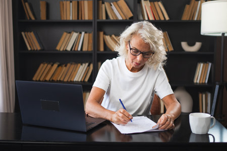 Middle aged senior woman using laptop computer writing notes at workplace. Focused mature old businesswoman work at office making written records doing online work. Mature boss professional workerの写真素材