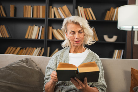 Comfort relaxed senior old elderly woman reading book at home. Mature woman resting enjoy calm day sitting on sofa at home alone. Adult lady spending free time keeping book smiling. Pension conceptの写真素材