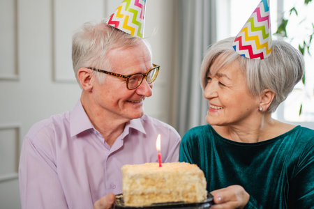 Make a wish. Family senior couple in party hat cap celebrating birthday anniversary together at home. Old man blowing out burning candles on birthday cake. Old woman wishes husband happy birthdayの写真素材