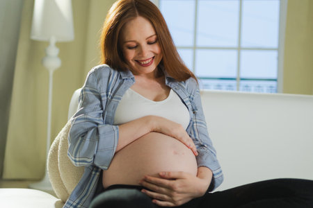 Pregnancy motherhood people expectation future. Pregnant woman touching big belly sitting on couch at home. Girl hugging her tummy enjoying pregnancy. Maternity tenderness parenthood new life conceptの写真素材