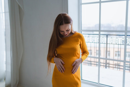 Pregnancy motherhood people expectation future. Pregnant woman with big belly standing near window at home. Girl hugging her tummy enjoying pregnancy. Maternity tenderness parenthood new life conceptの写真素材