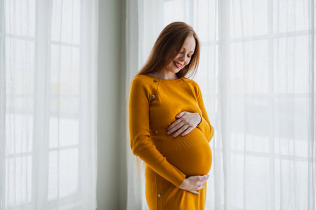 Pregnancy motherhood people expectation future. Pregnant woman with big belly standing near window at home. Girl hugging her tummy enjoying pregnancy. Maternity tenderness parenthood new life conceptの写真素材