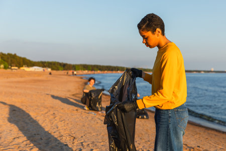 Earth day. Volunteers activists team collects garbage cleaning of beach coastal zone. Woman puts plastic bottle trash in garbage bag on ocean shore. Environmental conservation coastal zone cleaningの写真素材