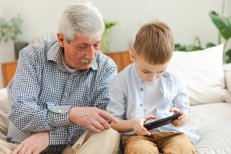 Happy family at home. Old senior man grandfather boy grandson enjoying time together watching video on phone. Good time at home. grandparent and child together. Older generation modern tech usageの写真素材