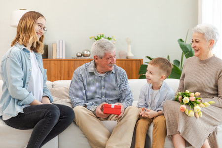 Happy family grandchildren gives gift box spring tulip flowers to grandparents. Smiling kids children with bouquet surprise gift box getting congratulations on grandparents day. Family celebratingの写真素材