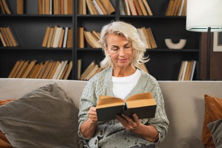 Comfort relaxed senior old elderly woman reading book at home. Mature woman resting enjoy calm day sitting on sofa at home alone. Adult lady spending free time keeping book smiling. Pension conceptの写真素材