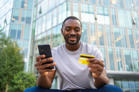 African man shopping online holding smartphone paying with gold credit card. Guy on urban street in...