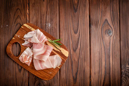 Raw bacon slices and rosemary sprig on wooden cutting board on wooden background. Top view. Copy space.の写真素材