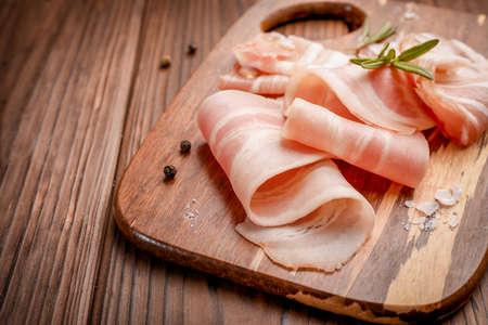 Raw bacon slices, salt and rosemary sprig on wooden cutting board on wooden background.の写真素材