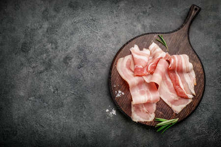 Raw bacon slices, salt and rosemary sprig on wooden cutting board on black background. Top view. Copy space.の写真素材