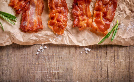 Fried crispy bacon slices, salt and rosemary sprigs on kraft paper on wooden background. Top view. Copy space.の写真素材