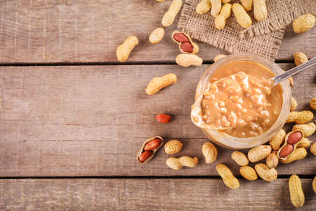 Glass jar full of crunchy peanut butter with peanuts in nutshell nearby on wooden background. Top...