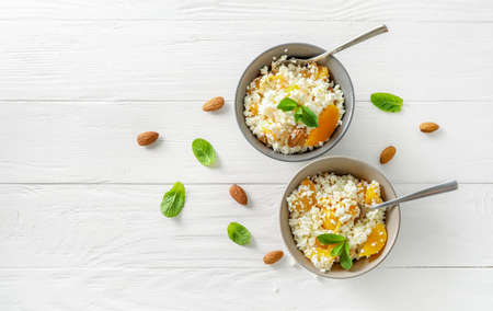 Fresh cottage cheese in bowls with mint leaves, peach pieces and almond nuts on white wooden...