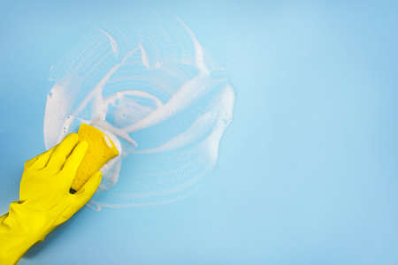 hand in a yellow rubber glove washes the wall with an orange soft rag, cleaning and washing the walls, blue background copy space, cleaning concept, spring cleaning, housework conceptの写真素材