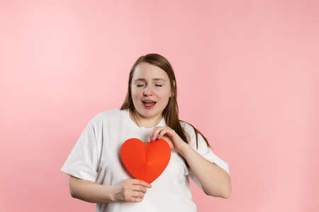 Enjoying young girl with brown hair in a white t-shirt on a pink background and red heart. High...