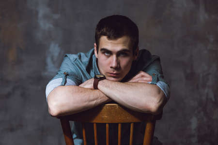Portrait. Young man with a short hair in a turquoise shirt sit on a chair and looking in the camera. High quality photoの写真素材