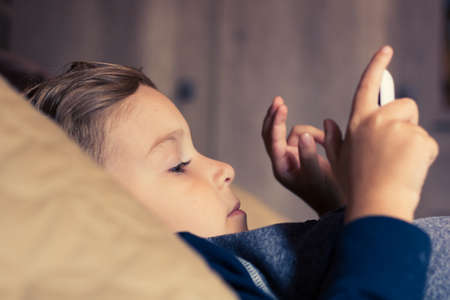 Little boy lying down on bed and watching cartoons on digital tablet in the evening.