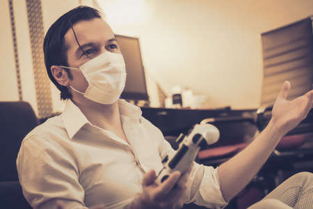 Man with protective face mask playing video games at home during a lockdown due to COVID-19 pandemic.の写真素材