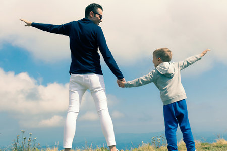 Back view of happy father and son holding hands and having fun against the sky at mountain peak.の写真素材