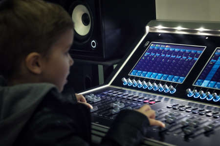 Kid learning to produce music and mixing sound on audio panel in recording studio.