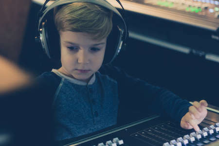 Cute kid with headphones editing music on audio mixer in recording studio.