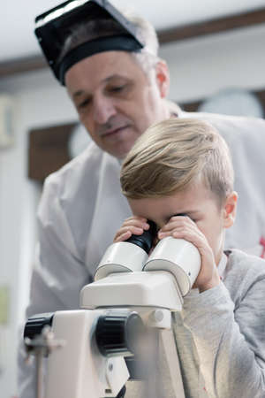 Small kid looking through microscope and examining something with help of science teacher in a laboratory.の写真素材