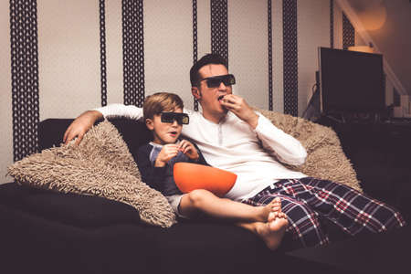Father and son watching movie with 3d glasses and eating popcorn while relaxing on the sofa.