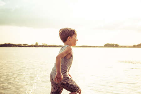 Happy boy having fun while running through water on the beach at sunset.の写真素材