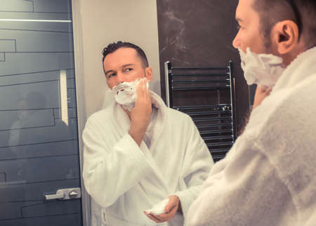 Reflection in a mirror of man applying shaving foam while shaving in the bathroom.の写真素材