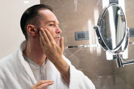 Man in bathrobe using face cream after shaving in the bathroom. The view is through the glass.の写真素材