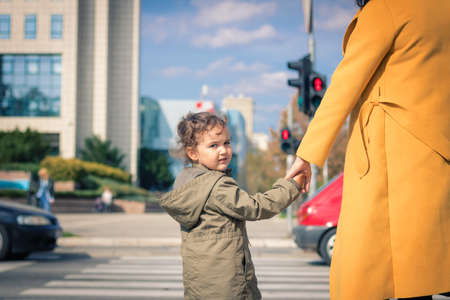 Small daughter holding mother's hand while waiting on zebra crossing in the city. Cute child crossing city street while holding mother's hand and looking at camera.の写真素材