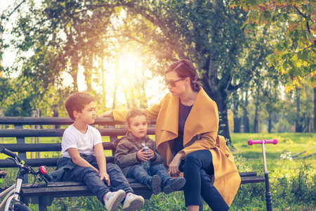 Little boy and girl relaxing on park bench with their mother. Young mother talking to her children while enjoying in day outdoors.の写真素材