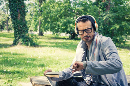 Worried man checking time on wristwatch while reading book and listening music on headphones in the park.の写真素材