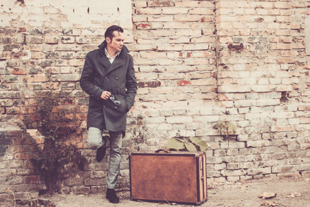 Full length of serious man in coat with vintage suitcase leaning on brick wall on contemplating.の写真素材
