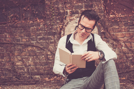 Styled man reading book while sitting outdoors against brick wall.の写真素材