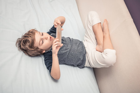 Little boy in pajama lying on bed and watching something online in the evening.の写真素材