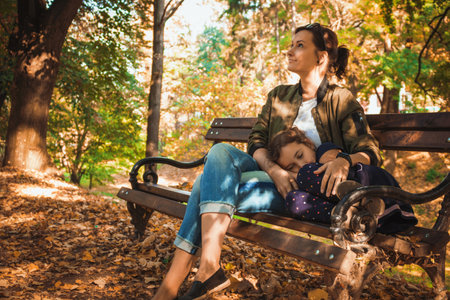 Mother and daughter enjoying in autumn day while relaxing on a bench in the park. Little girl is napping in mother's lap.の写真素材
