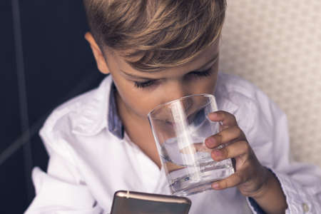 Kind drinking water while watching cartoons on smart phone. Little boy refreshing himself and using...