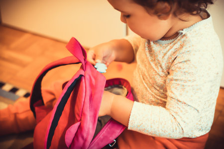 Small girl sitting on the floor and packing her backpack while preparing for first day at kindergarten.の写真素材