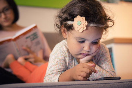 Cute little girl using mobile phone and playing games while her mother is reading book in the...