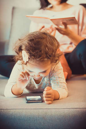Cute small girl surfing the net on smart phone while relaxing at home with her mother.の写真素材