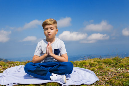 Calm boy with hands clasped practicing Yoga with eyes closed on top of the mountain. Serene kid in namaste pose against the sky.の写真素材